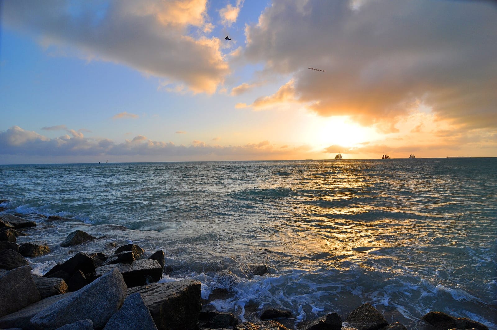 Truman Beach: sunset over ocean with rocks, waves, sailboats on horizon